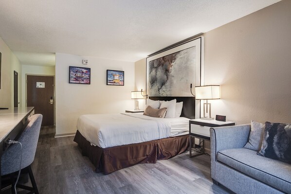 Quarto de hotel com cama, escrivaninha e cadeira, um quadro abstrato grande na parede e duas fotos emolduradas.
