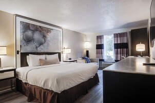 Quarto de hotel com uma cama grande, mesas de cabeceira, uma escrivaninha e um quadro abstrato grande na parede.