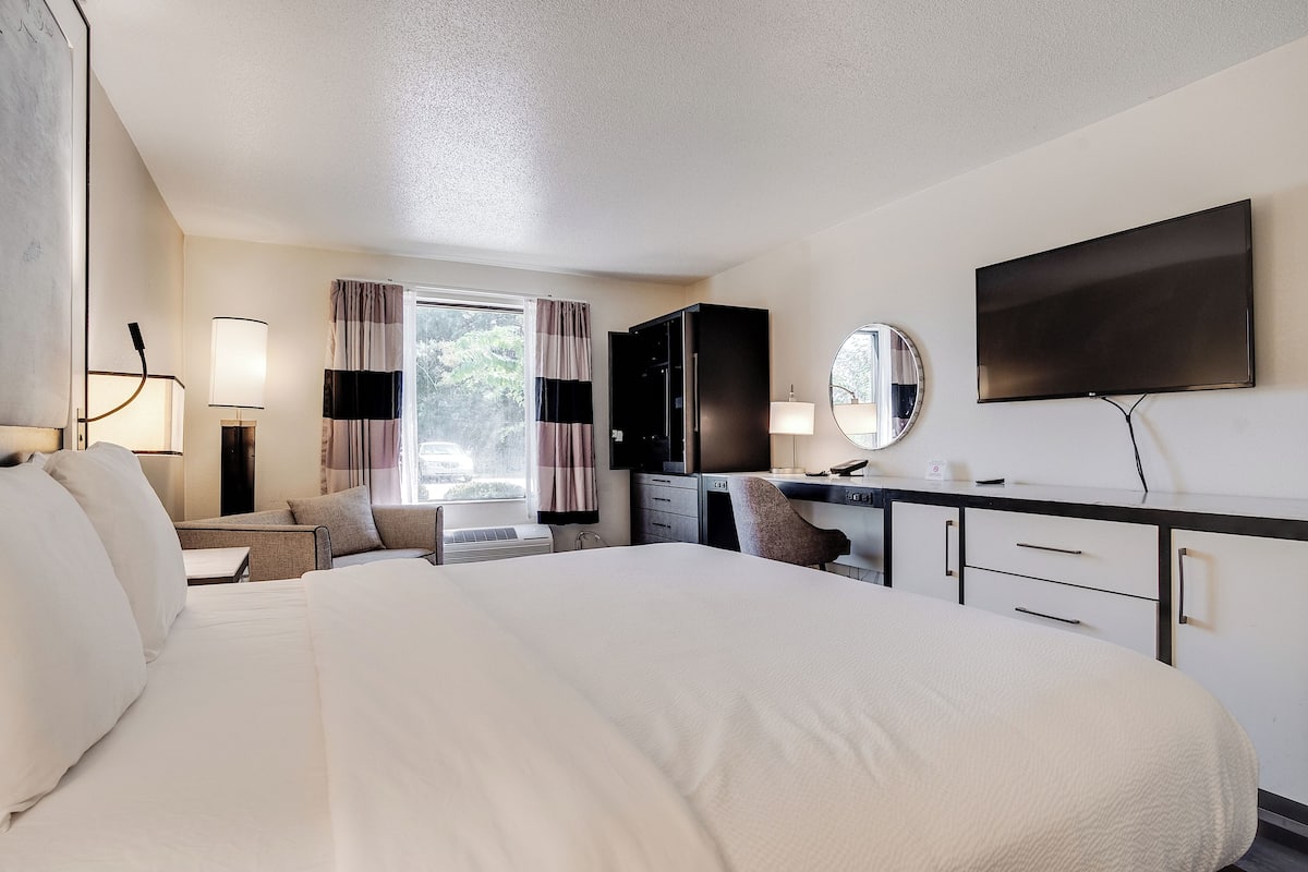 A hotel room with a large bed, a desk with a computer, a TV, and a window with curtains.