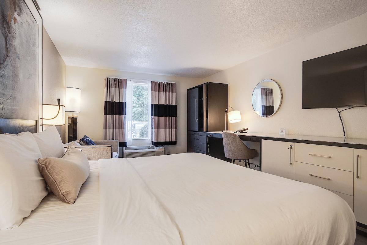A hotel room with a large bed, a desk with a chair, a flat-screen TV, a mirror, and a window with curtains.