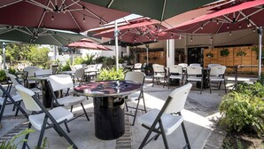 Terrassa exterior amb taules i cadires sota grans ombrel·les, envoltada de vegetació i un edifici al fons.