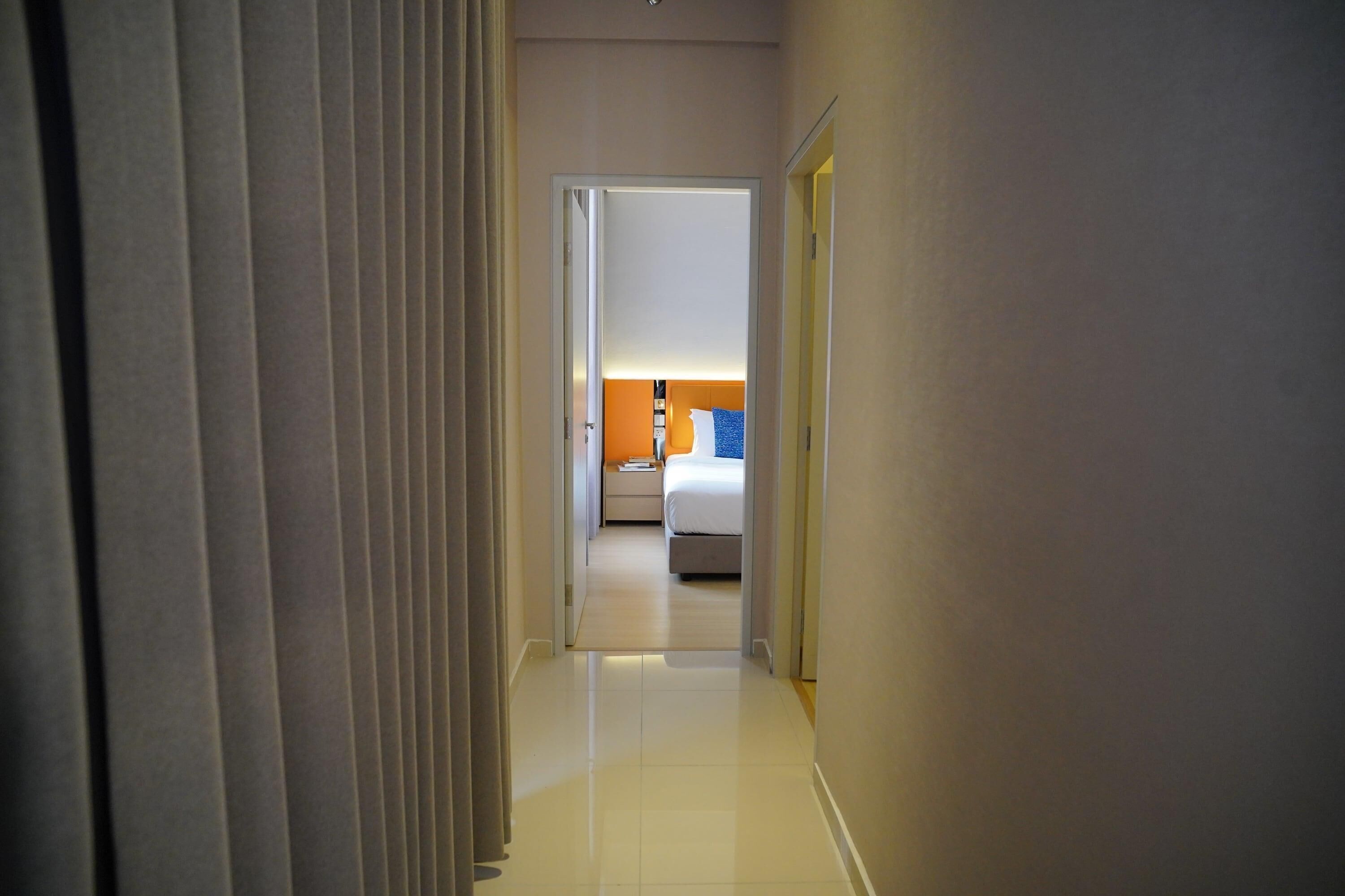 A hotel hallway leading to a bedroom with a bed, nightstand, and lamp.