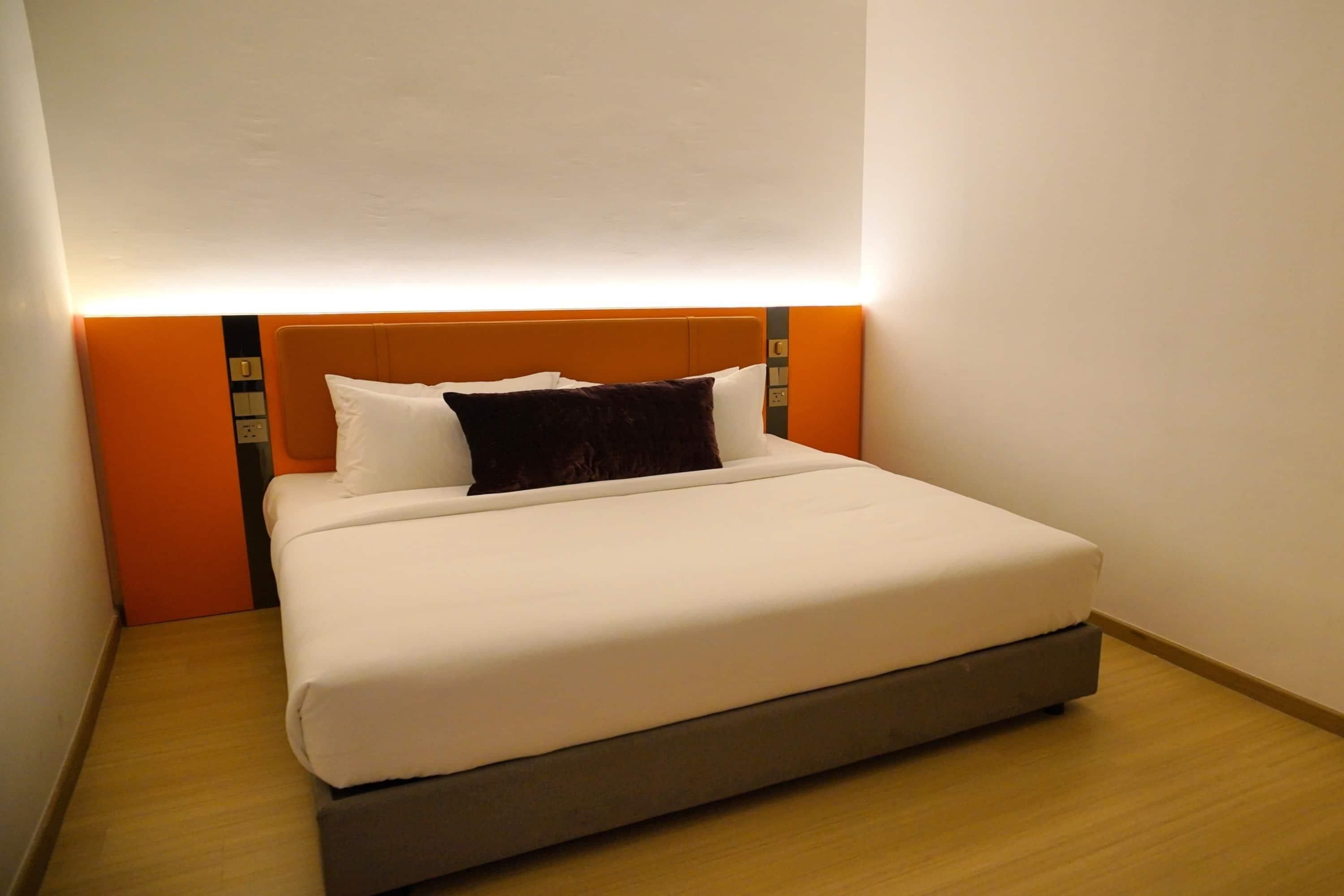 A neatly made bed with white linens and a dark brown pillow, a wooden floor, and a wall with a light switch and a thermostat.