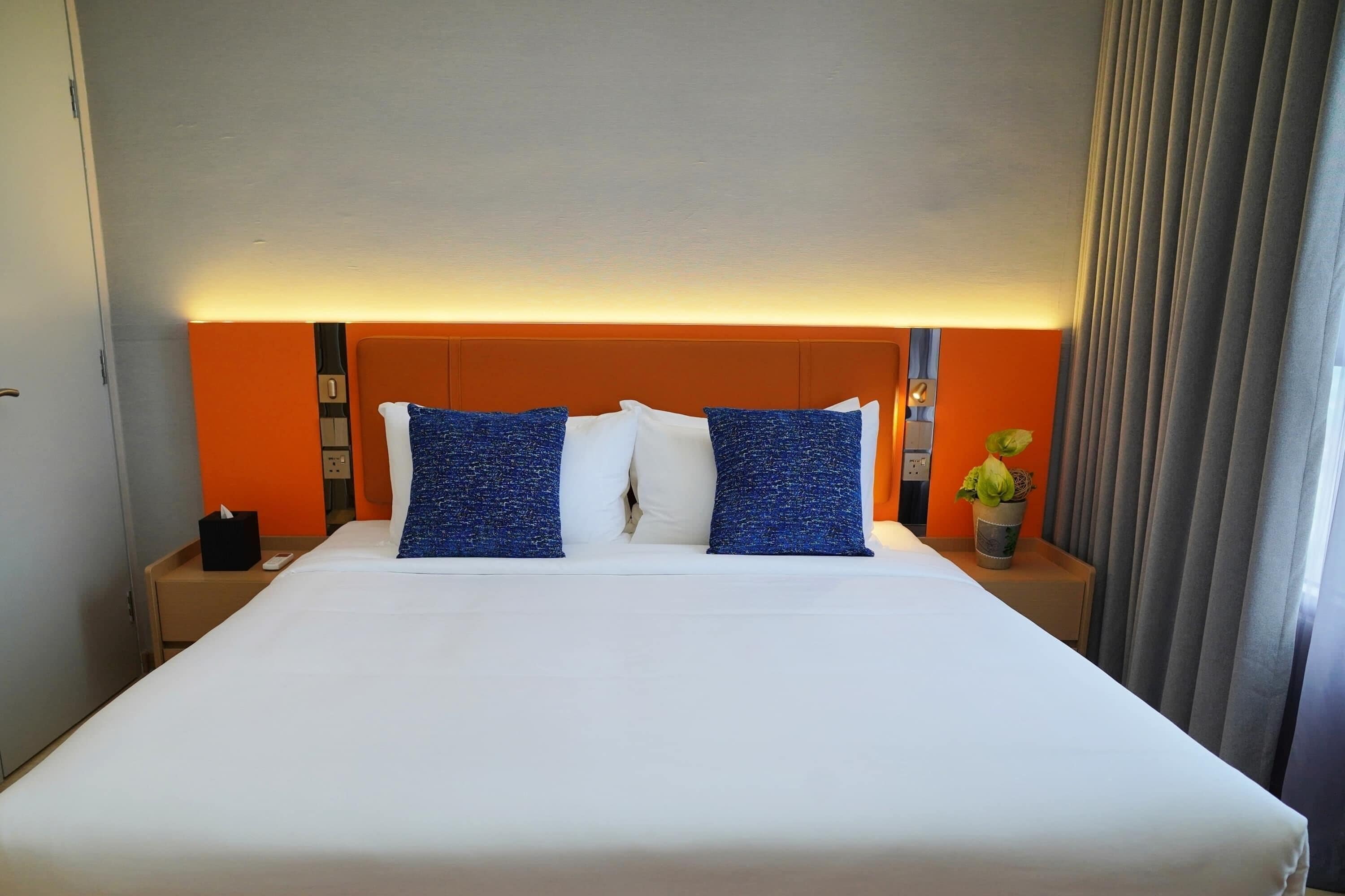 Una camera d'albergo con un letto grande, una testiera arancione, biancheria da letto bianca e un comodino con una pianta.