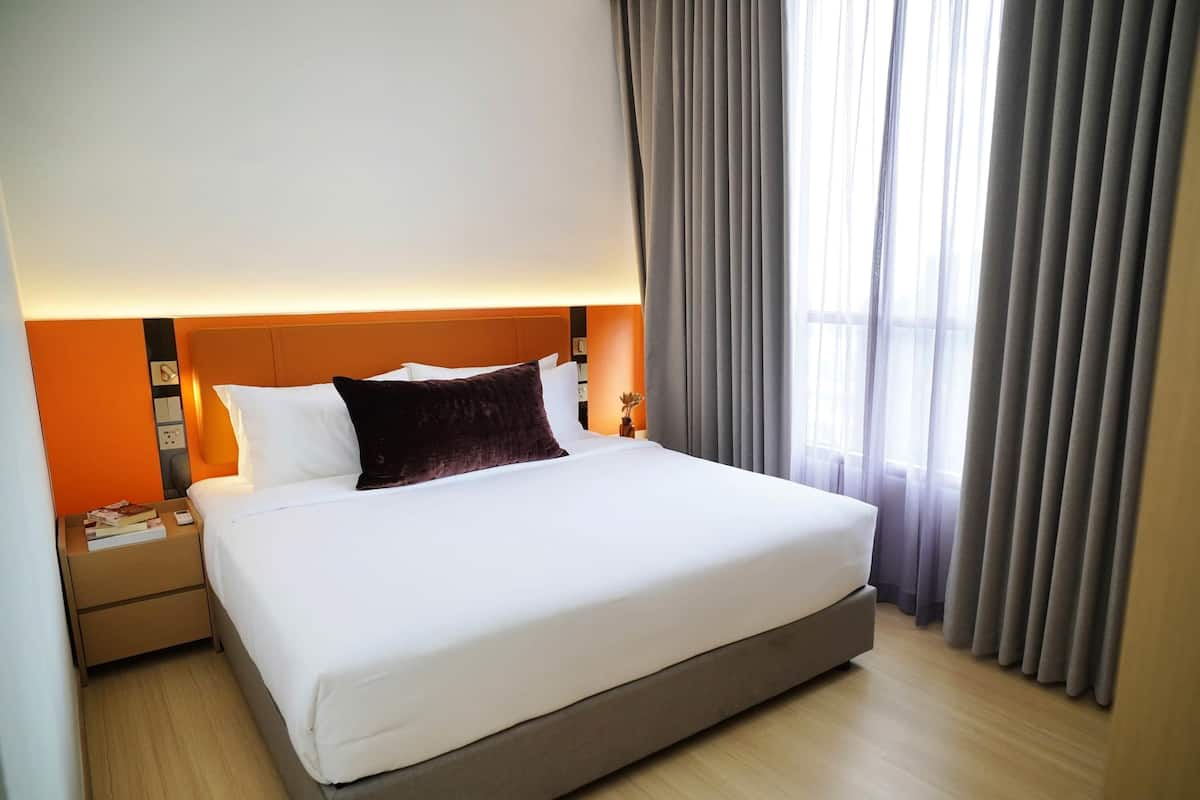 Un lit bien fait, avec du linge de lit blanc et un coussin décoratif marron foncé, une table de chevet en bois avec un livre et une télécommande, et une grande fenêtre avec des rideaux gris.