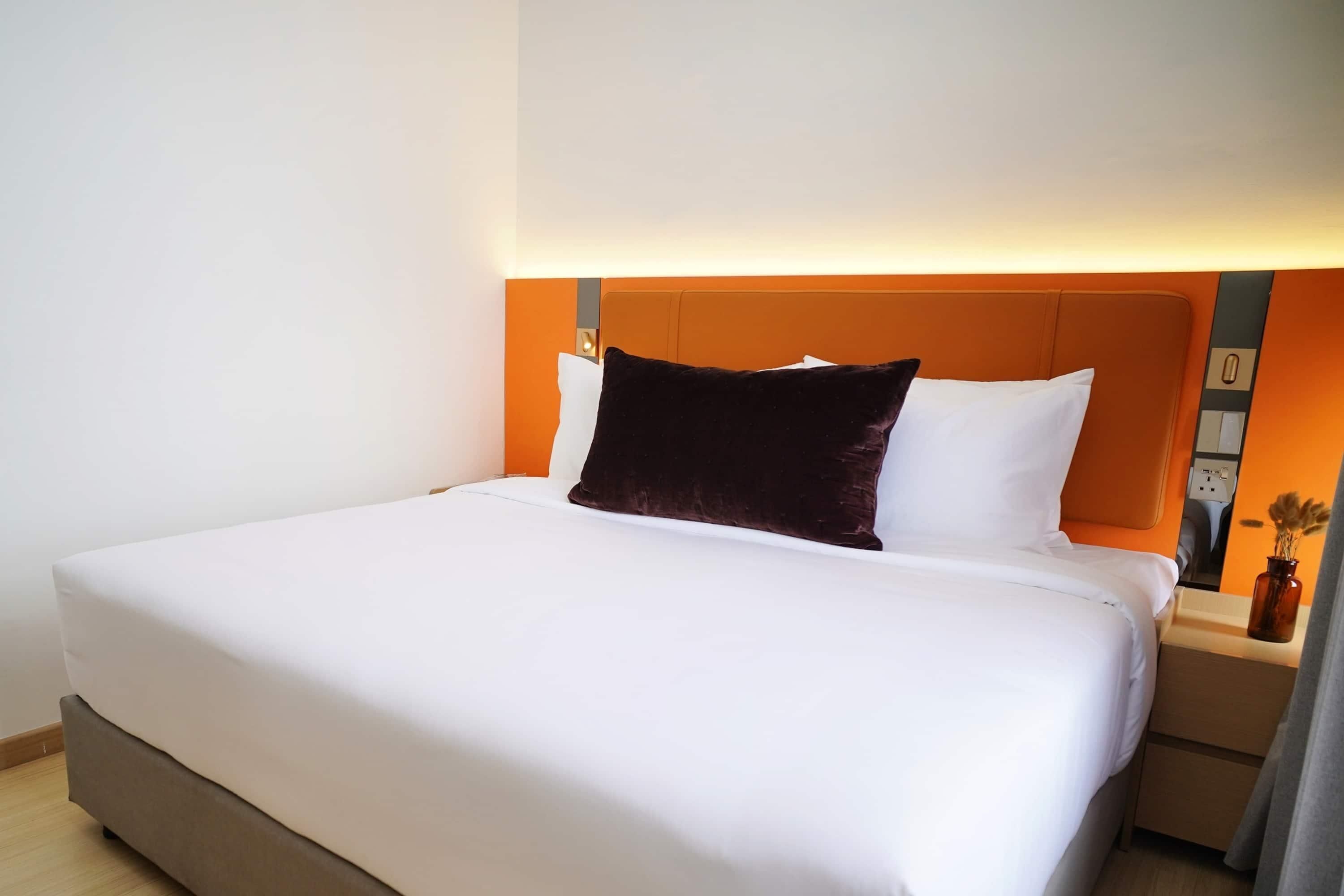 A neatly made bed with white linens and a dark brown pillow, a bedside table with a vase, and a wall-mounted light fixture.