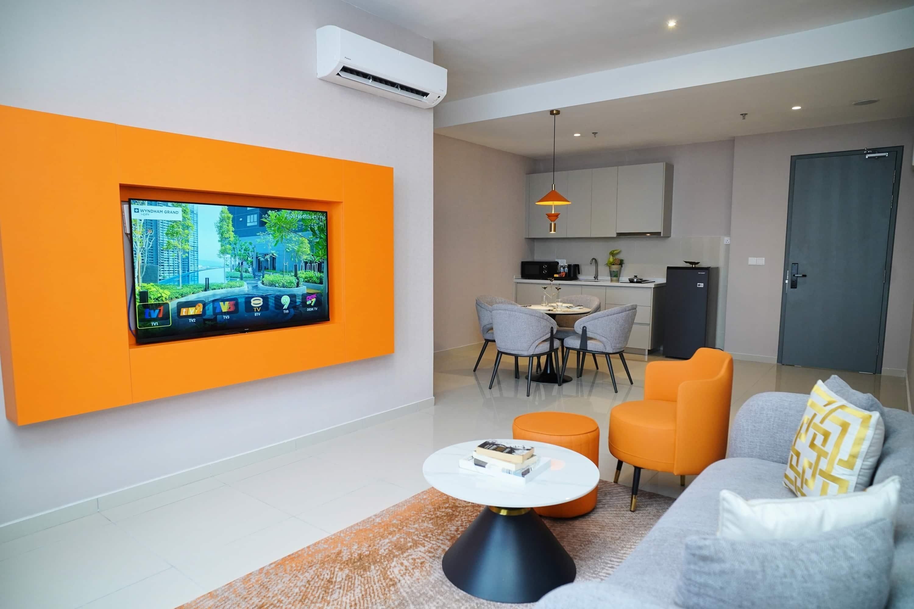 Una sala de estar moderna con un televisor de pantalla plana montado en una pared naranja, un sofá gris con cojines amarillos y una pequeña mesa de centro redonda.