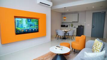 Una sala de estar moderna con un televisor de pantalla plana montado en una pared naranja, un sofá gris con cojines amarillos y una pequeña mesa de centro redonda.
