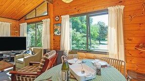 L'intérieur d'un chalet en bois comprend une table à manger pour quatre personnes, un canapé, un téléviseur et de grandes fenêtres avec des rideaux.