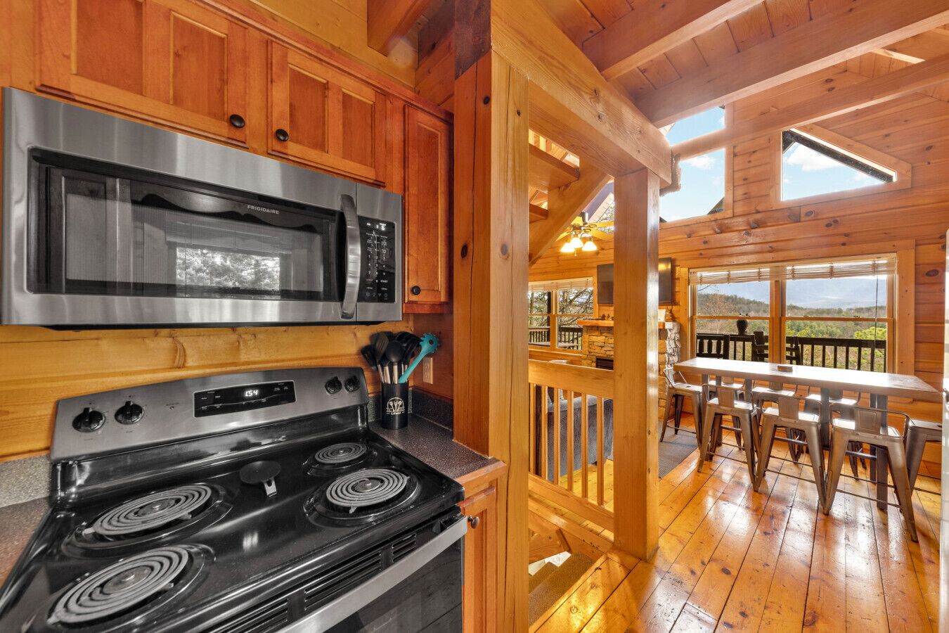 A kitchen with a stainless steel microwave and stove, wooden cabinets, and a view of the outdoor deck with a table and chairs.