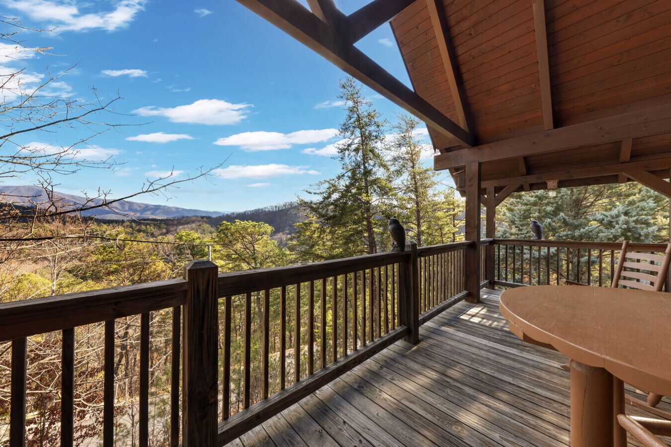 A wooden deck with a table and chairs, overlooking a mountainous landscape with trees and a clear sky.