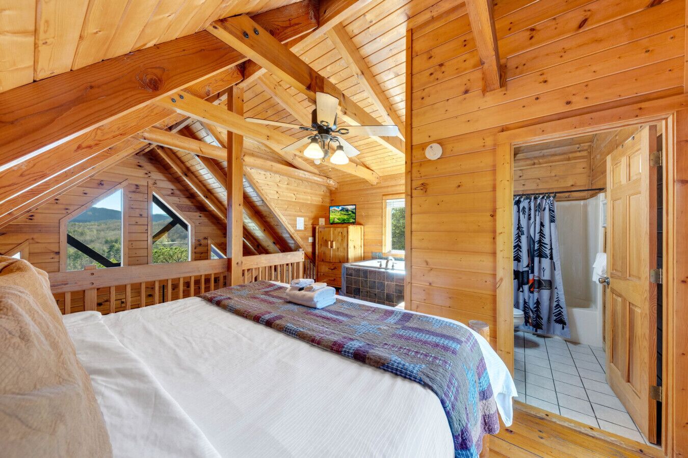 A wooden cabin bedroom with a bed, a ceiling fan, a television, and a bathroom with a shower curtain.