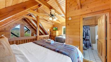A wooden cabin bedroom with a bed, a ceiling fan, a television, and a bathroom with a shower curtain.