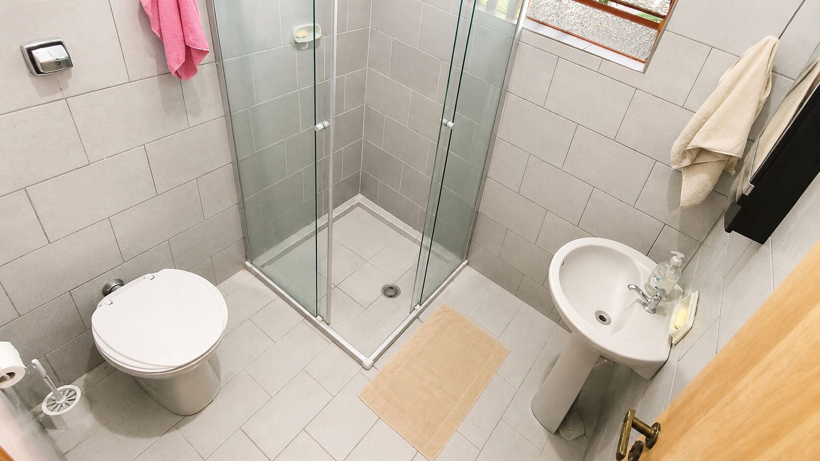 Ein modernes Badezimmer mit einer Duschkabine aus Glas, Toilette, Waschbecken mit Seifenspender und einer beige farbenen Badmatte.