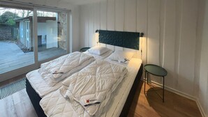 Un lit double avec une literie blanche et une tête de lit, une petite table à côté, et une vue sur un jardin par une porte coulissante.