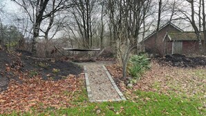 Un jardin avec un sentier en bois, un sol recouvert de feuilles et un bâtiment en briques en arrière-plan.