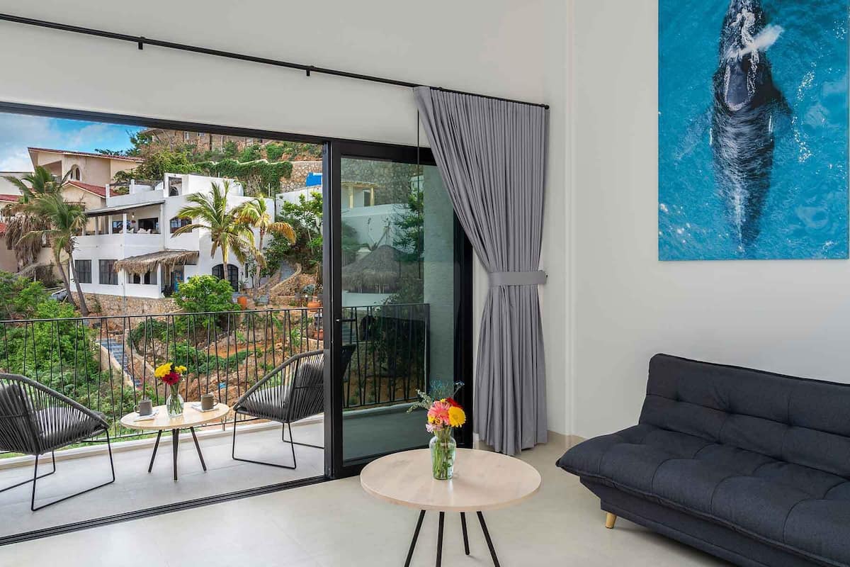 Un salon moderne avec un balcon, un canapé, une table ronde ornée d’un vase de fleurs et une grande fenêtre donnant sur un bâtiment blanc et une végétation luxuriante.