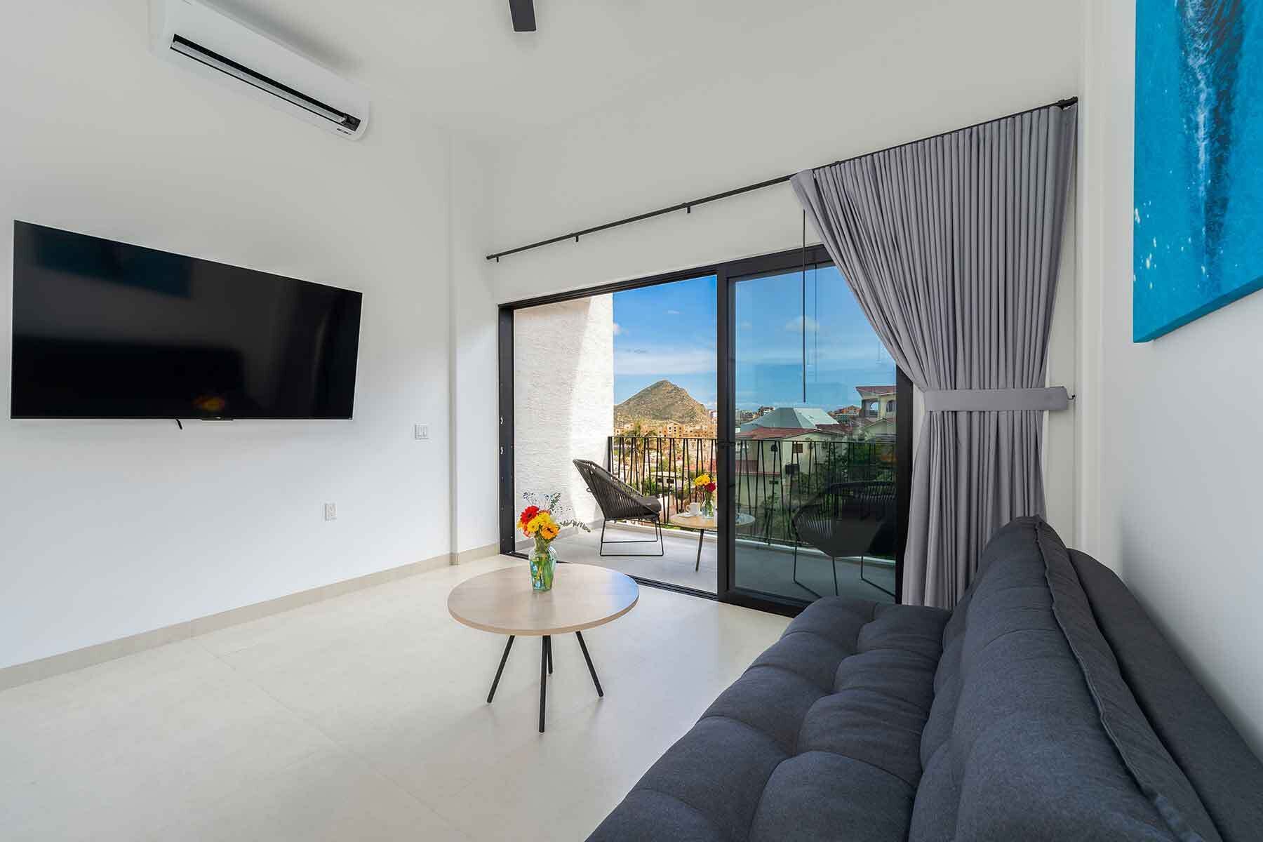Un salon moderne avec un téléviseur à écran plat, un canapé gris, une table basse ronde ornée d’un vase de fleurs et un balcon offrant une vue sur les bâtiments et une montagne.