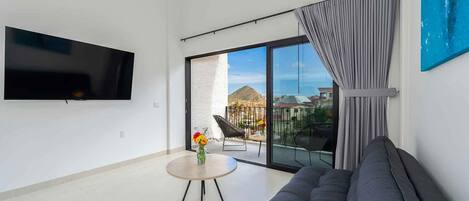 Un salon moderne avec un téléviseur à écran plat, un canapé gris, une table basse ronde ornée d’un vase de fleurs et un balcon offrant une vue sur les bâtiments et une montagne.