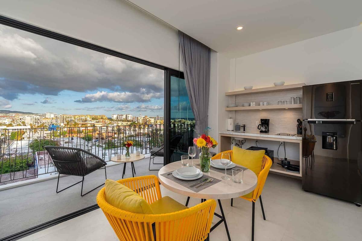 Un appartement moderne doté d’un balcon, d’un coin repas avec des chaises jaunes, d’une cuisine avec un réfrigérateur en acier inoxydable et offrant une vue sur la ville.