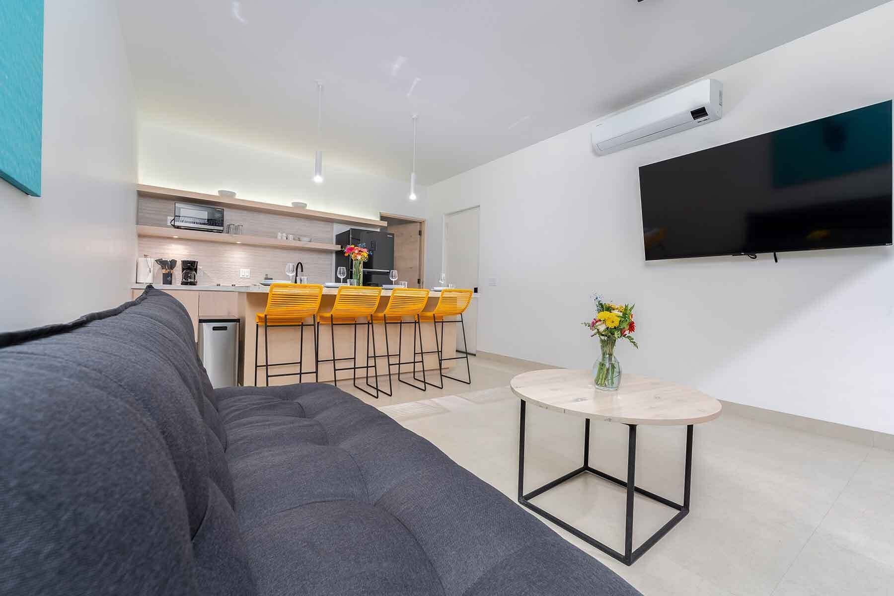 A modern living room with a grey sofa, a small round table with a vase, and a kitchen area with wooden cabinets and yellow chairs.
