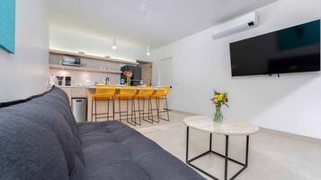 A modern living room with a grey sofa, a small round table with a vase, and a kitchen area with wooden cabinets and yellow chairs.