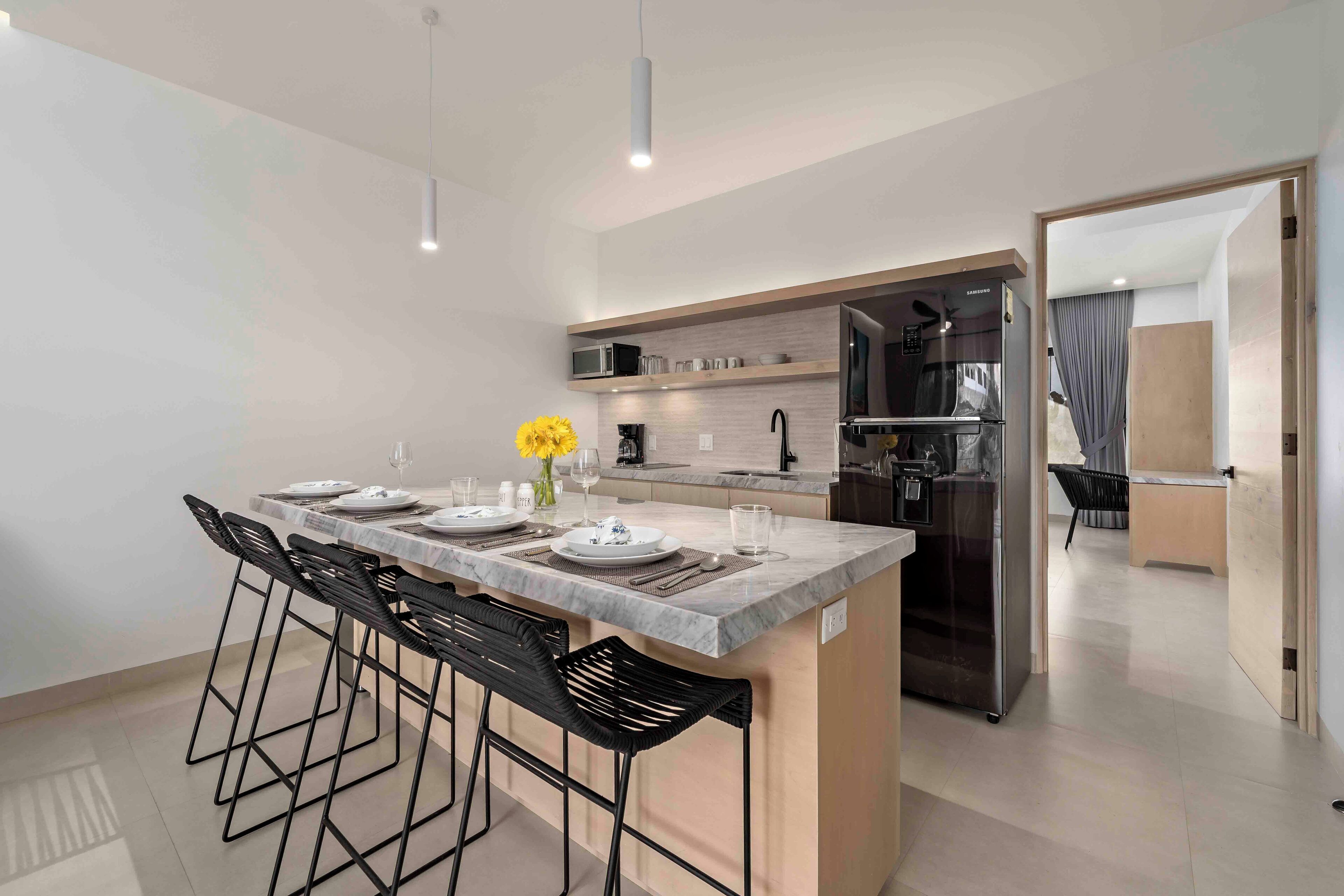 A modern kitchen with a marble countertop, black bar stools, a built-in microwave, and a stainless steel refrigerator.