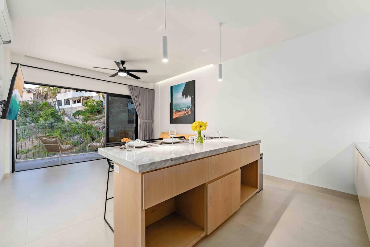 Une cuisine moderne dotée d’un grand îlot central, de meubles en bois et offrant une vue sur un balcon aménagé avec un espace salon extérieur.