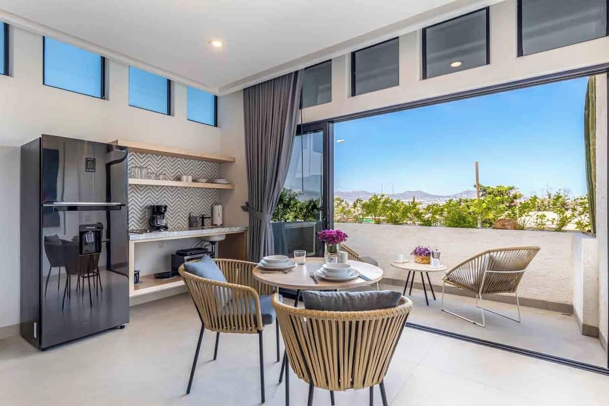 Une cuisine moderne dotée d’un réfrigérateur encastré, un coin repas avec des chaises en osier et une grande porte-fenêtre coulissante donnant sur un balcon avec vue sur le paysage.