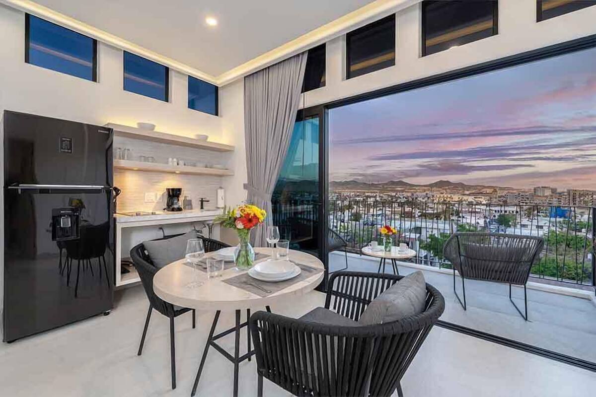 Une cuisine moderne équipée d’un réfrigérateur, d’une table à manger et de chaises. Un balcon offre une vue sur le paysage urbain.