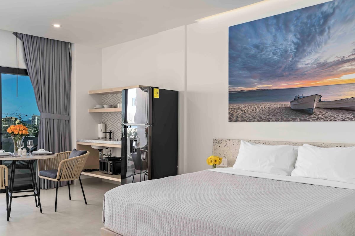 Une chambre d’hôtel moderne comprenant un lit, un coin repas avec des chaises, un réfrigérateur et un tableau représentant un coucher de soleil sur l’océan.