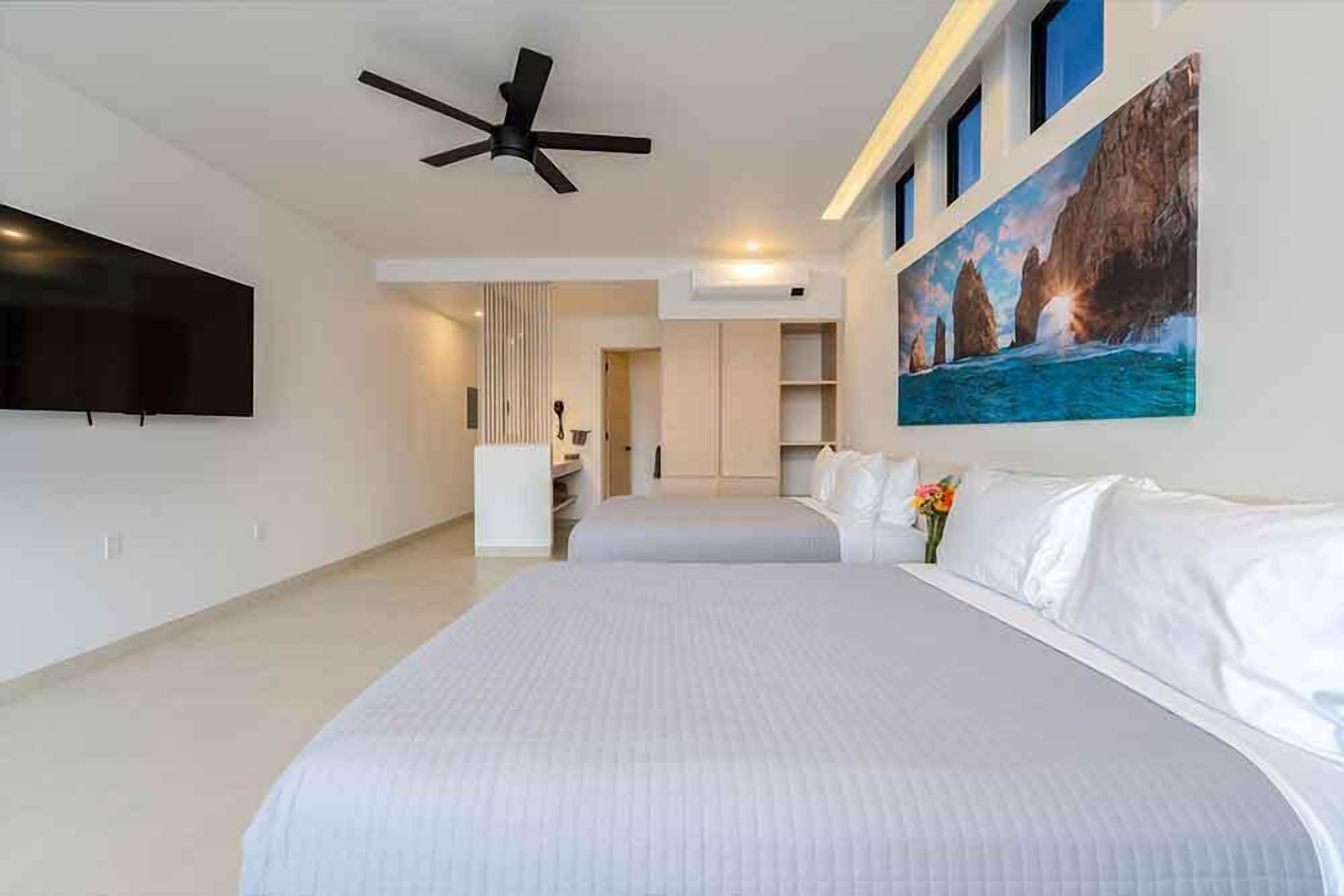 Une chambre d’hôtel avec un grand lit, un ventilateur de plafond, un téléviseur à écran plat et un tableau représentant un paysage côtier.