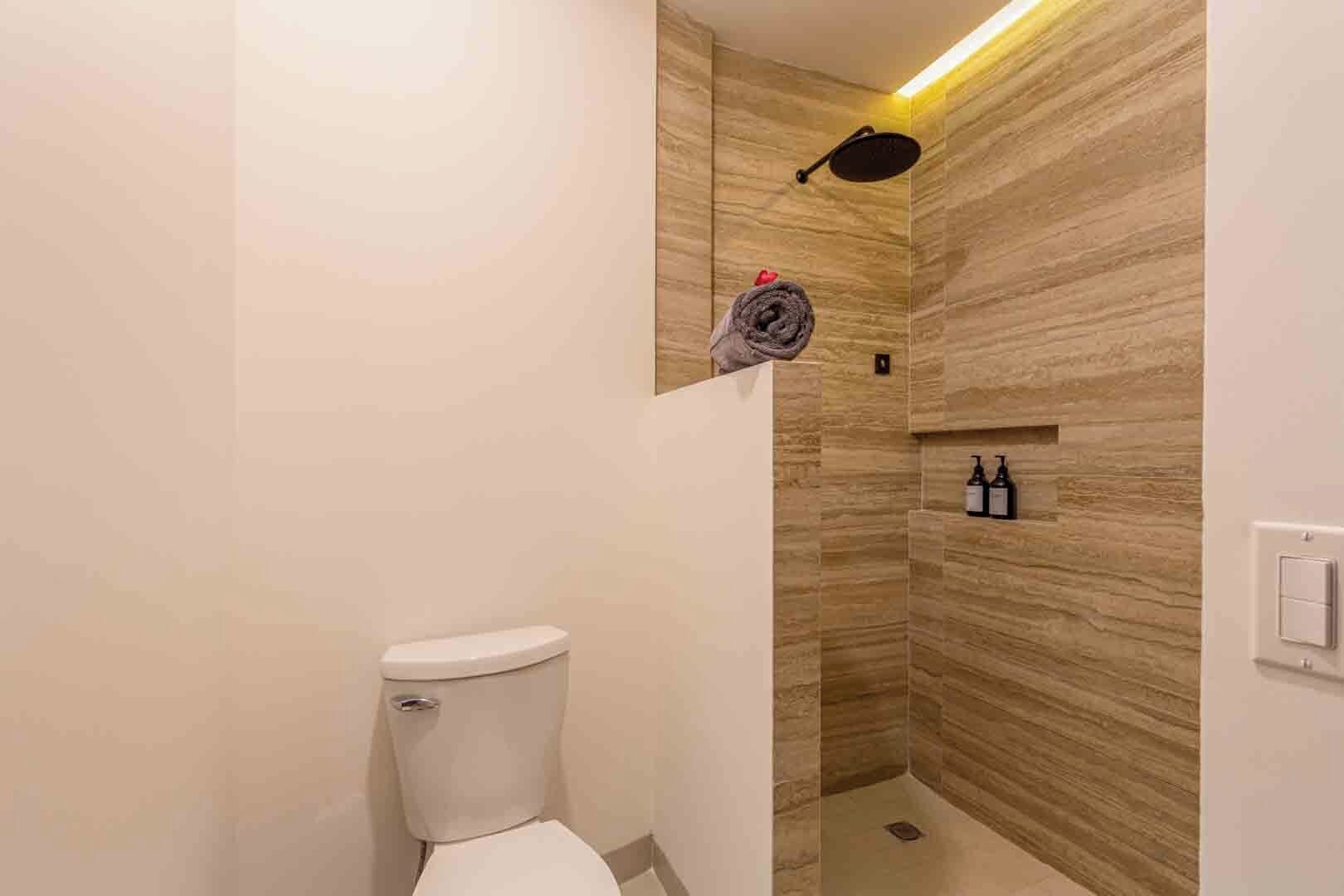 A modern bathroom with a toilet, a shower with a wooden wall, and a shelf with bottles.
