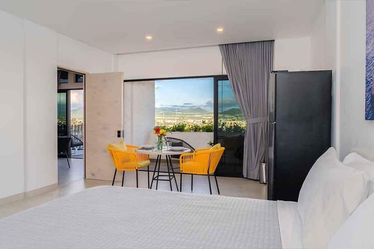 Une chambre d’hôtel moderne avec un grand lit, un coin repas avec des chaises jaunes, une vue sur le paysage et un réfrigérateur noir.