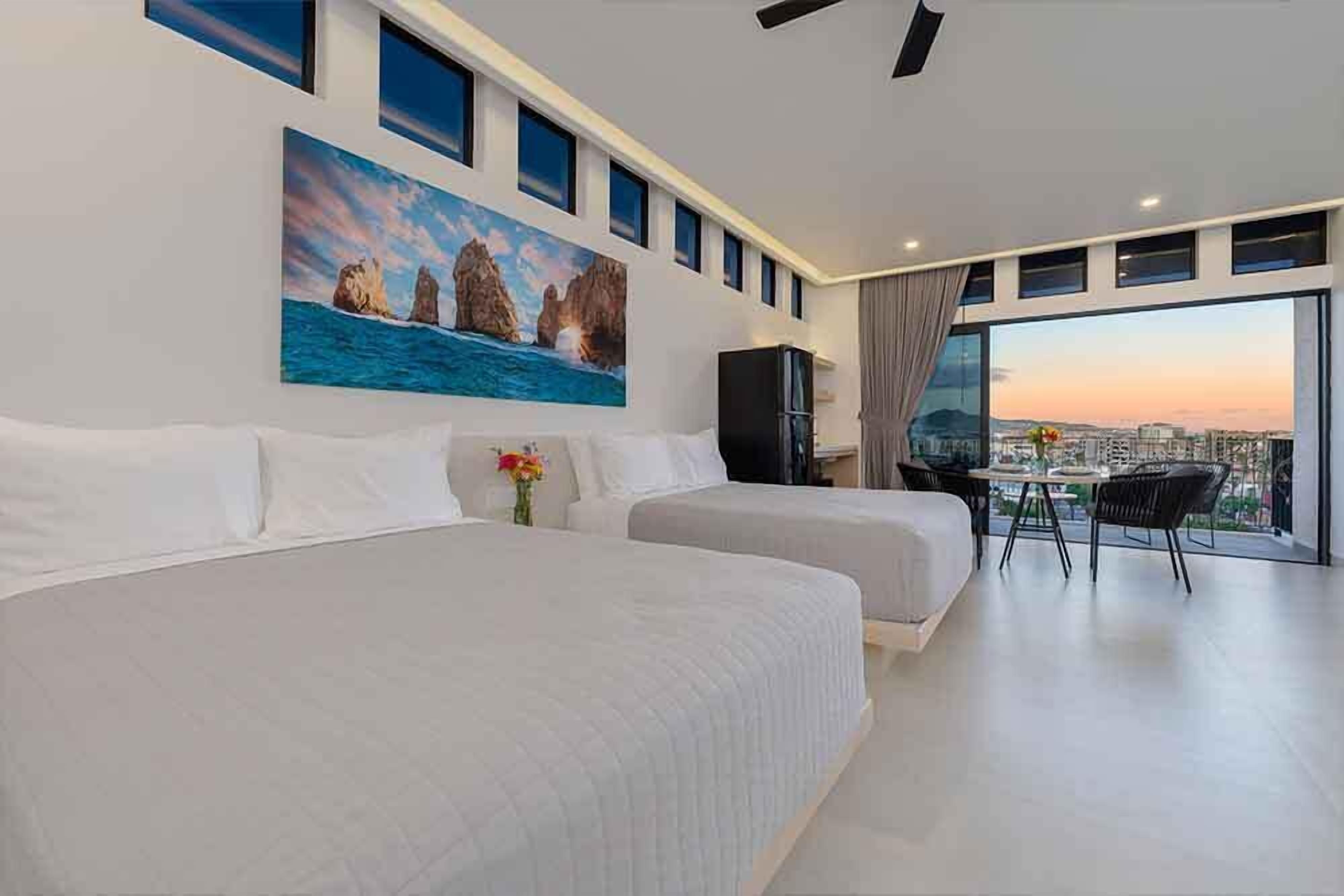 A hotel room with two beds, a painting of a coastal scene, a desk with a chair, and a large window offering a city view.