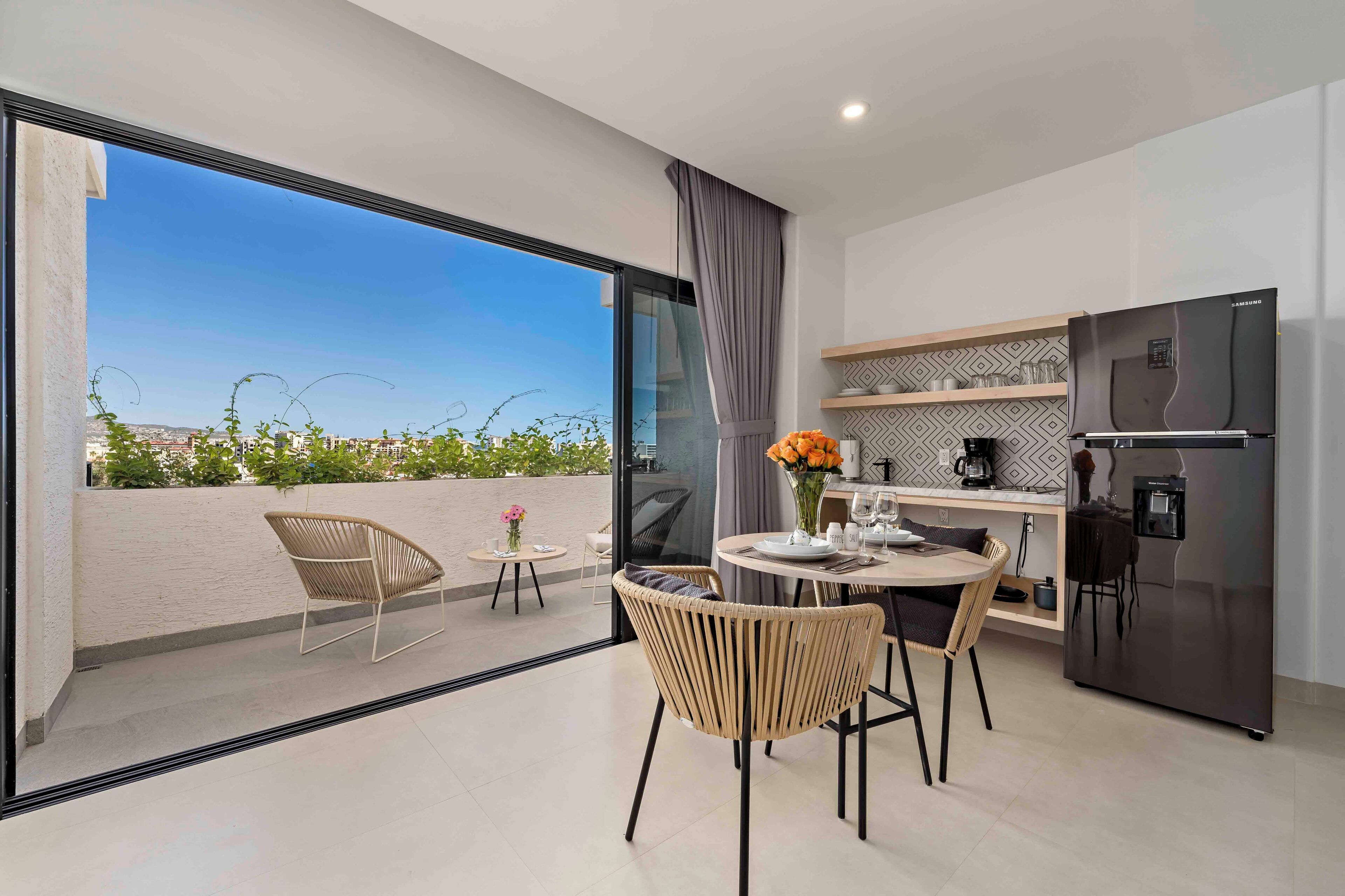 A modern kitchen with a large refrigerator, dining table, and chairs. A balcony with a view of the city is visible through the glass door.