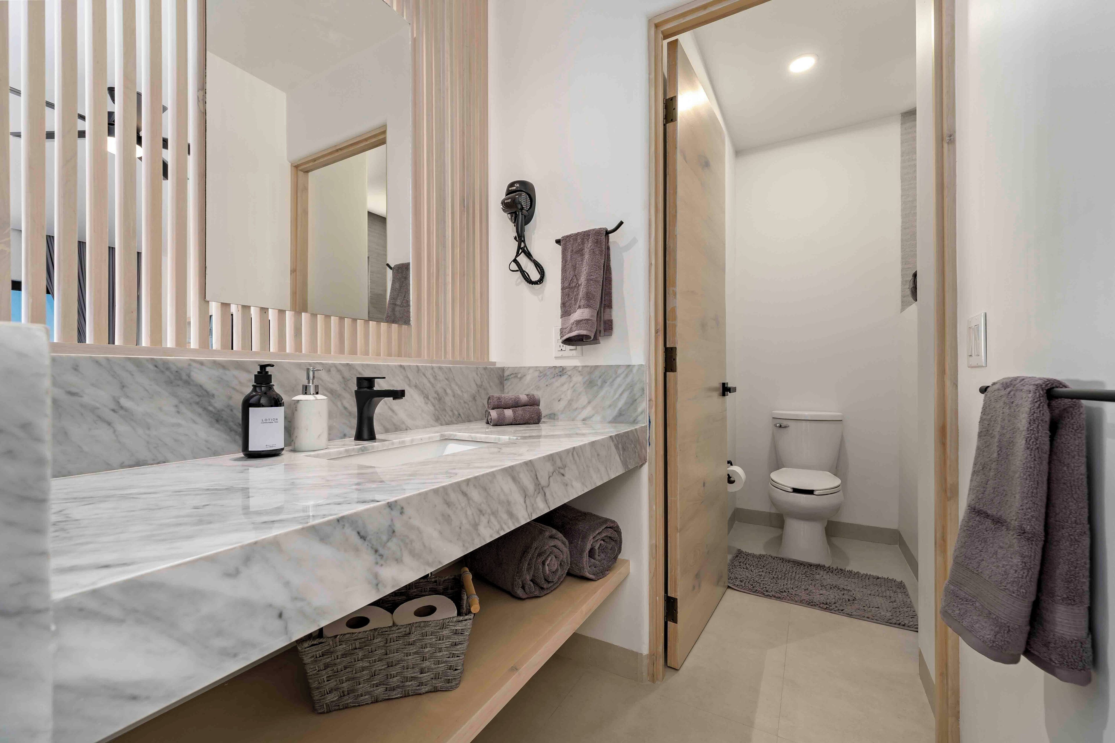 Un baño moderno con encimera de mármol, un secador de pelo montado en la pared y un inodoro visible a través de una puerta abierta.