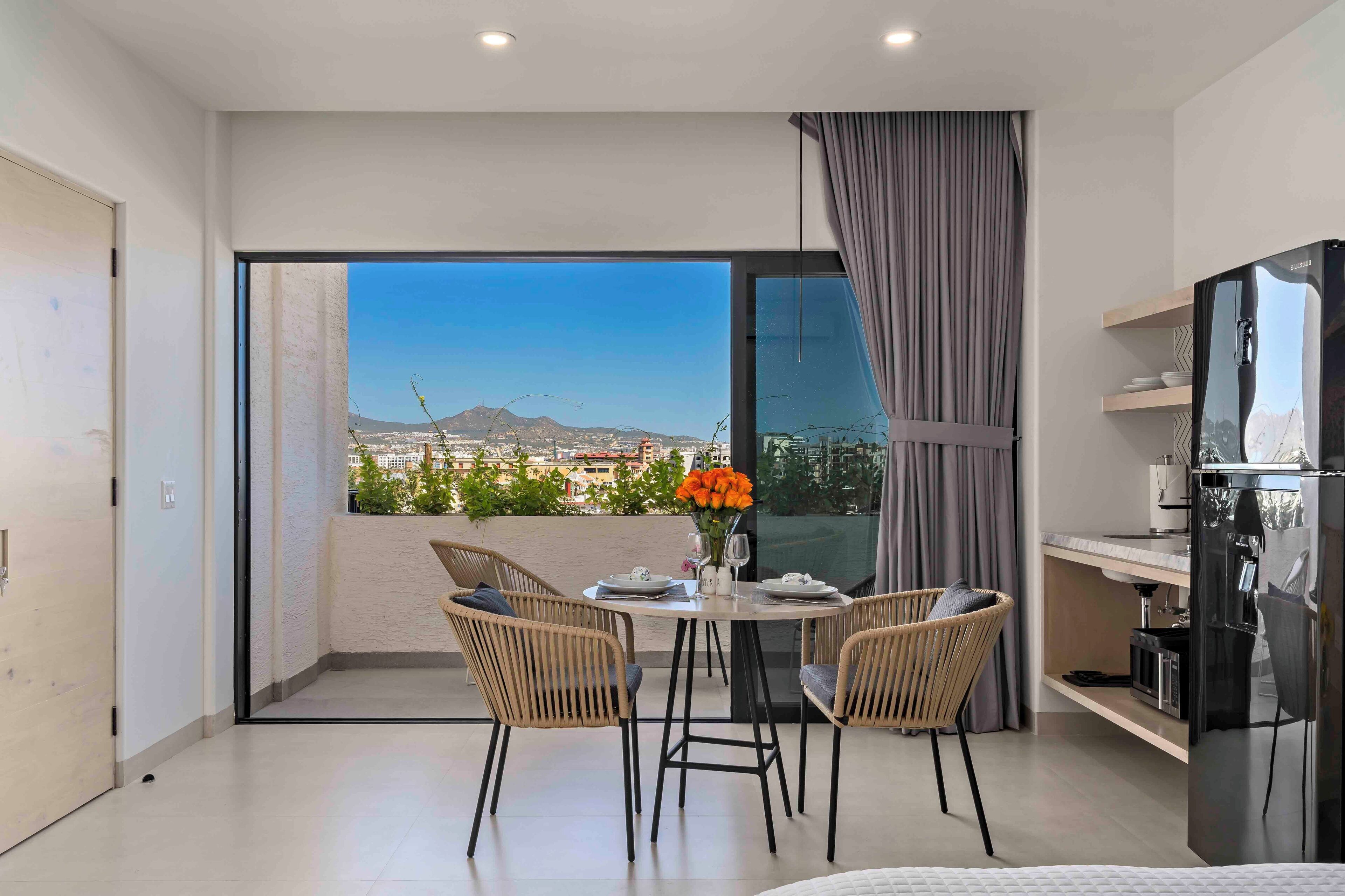 A modern room with a balcony, a dining table with chairs, a refrigerator, and a view of a cityscape.