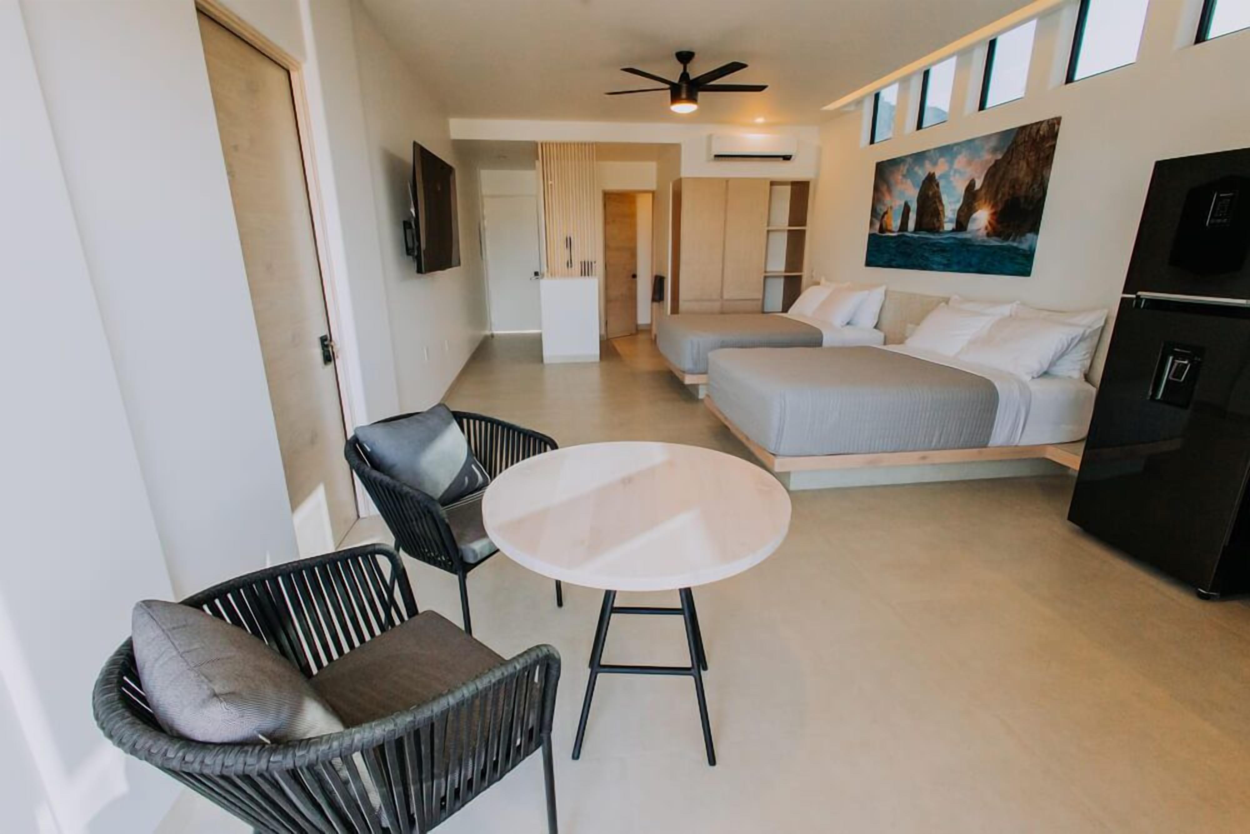 Quarto de hotel compacto com cama, duas mesas de cabeceira, uma cadeira, uma mesinha redonda, um ventilador de teto e um quadro com uma paisagem costeira.