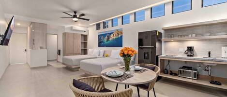 Una habitación de hotel moderna con una cama, una zona de comedor con mesa y sillas, una cocina con refrigerador y microondas, y un amplio ventanal con vista al mar.