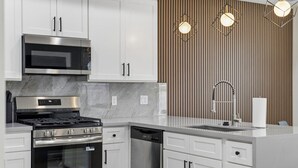 Una cocina moderna con armarios blancos, electrodomésticos de acero inoxidable, microondas, estufa y lavavajillas. Hay tres lámparas colgantes sobre el fregadero.
