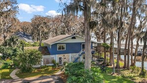 A single-story house with a blue exterior and a white porch, surrounded by lush greenery and a body of water. - Lake Eustis Waterfront Sunset House 3/2 Amazing Views! Boat Dock Available!  (Eustis)