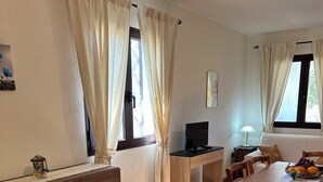 A cozy room with a wooden desk, a television, and a dining table with chairs. There are curtains on the windows, and a lamp on the side table. - DOMO on NEULACHE Country villa consisting of two apts. 5 minutes from the sea (Sardegna)
