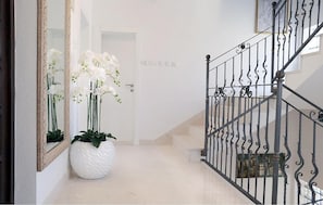 A hallway with a white floor, a large white vase with white flowers, a mirror, and a black wrought iron staircase. - Lovely home in Cilipi (Cilipi)
