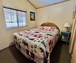 Ein Schlafzimmer mit Bett, einem Fenster mit Jalousien, einem Wandbild und einem kleinen Tisch mit Lampe.