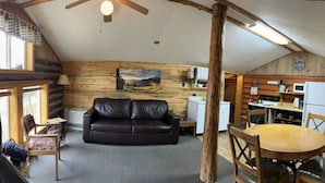 Et hyggeligt sommerhusinteriør med lædersofa, trævægge, spisebord og et køkken.