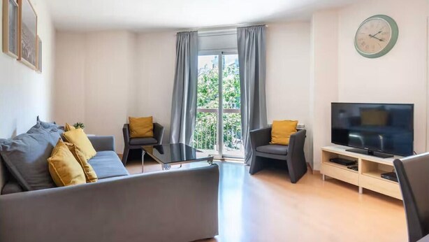 Una sala moderna con un sofá, dos sillones, una mesa de centro, un televisor y un balcón con cortinas.