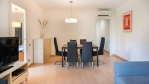 Una sala moderna con una televisión de pantalla plana, una mesa de comedor con sillas, un sofá, un armario blanco y una lámpara colgante.