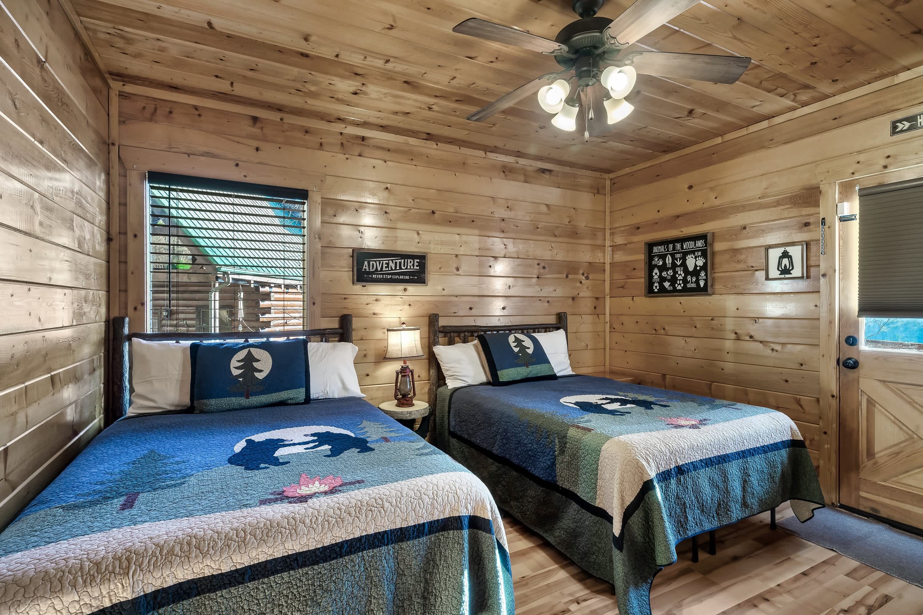 A wooden cabin room with two beds, a ceiling fan, and a window with blinds.