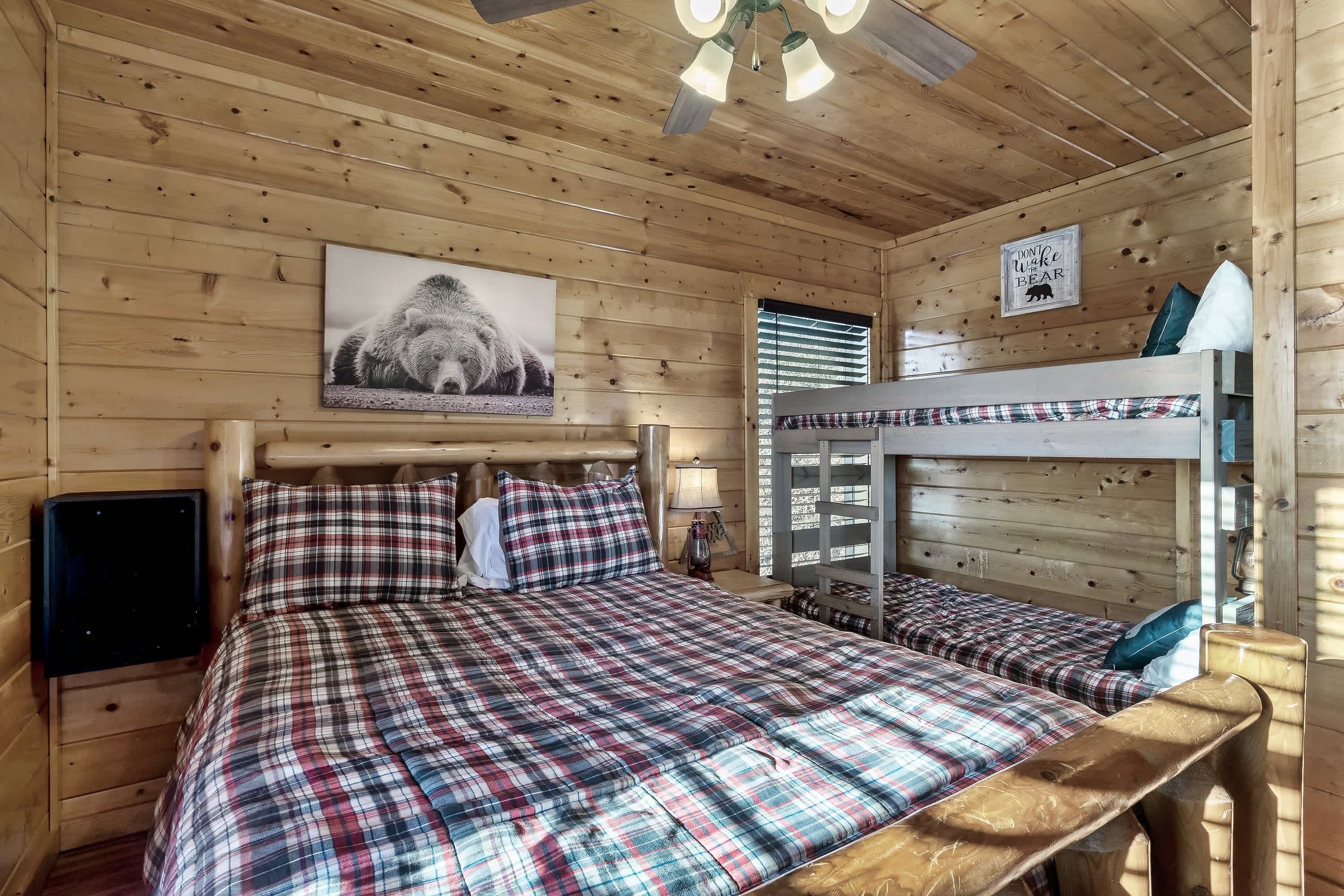 A wooden cabin room with a bunk bed, a large bed with plaid bedding, a mounted picture of a bear, and a sign that reads "Into the Woods Bear".